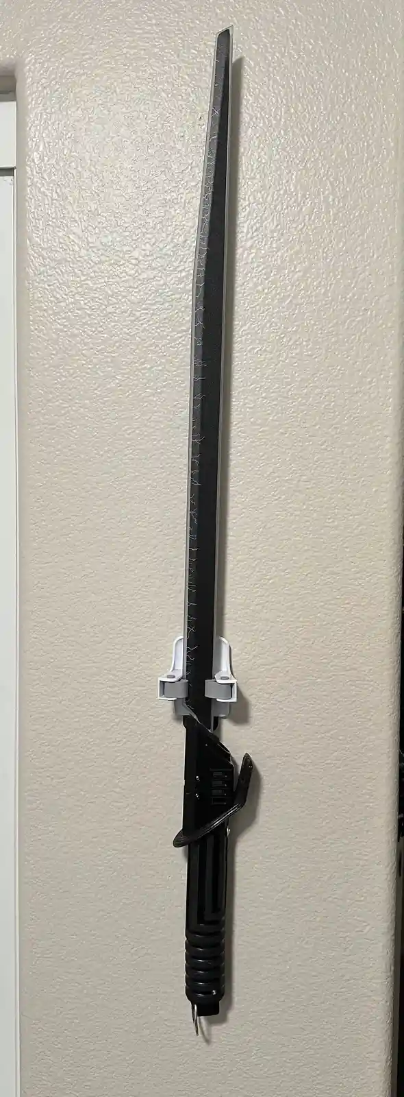 A black lightsaber hilt with a blade is mounted vertically on a beige textured wall using two white plastic clips. The blade…
