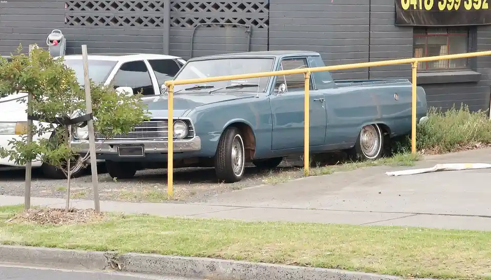 A light blue vintage pickup truck is parked in a lot next to a yellow railing, with its front wheel missing and the fender…