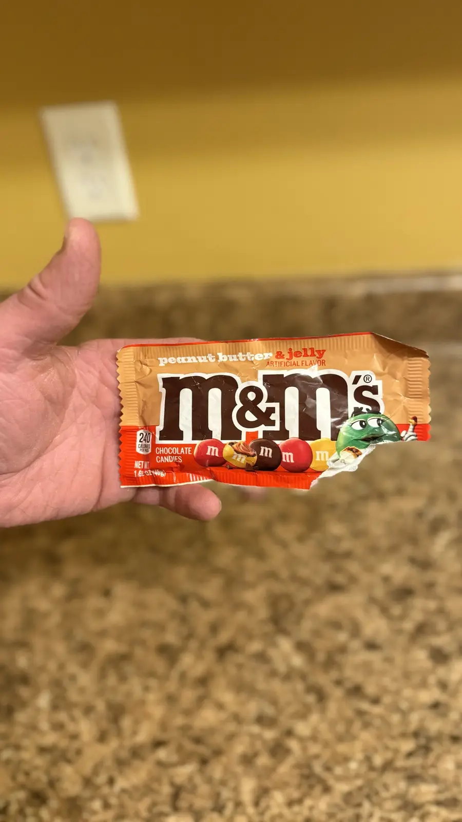 A hand holds a partially opened, torn package of M&M's peanut butter & jelly flavor chocolate candies. The wrapper shows the…