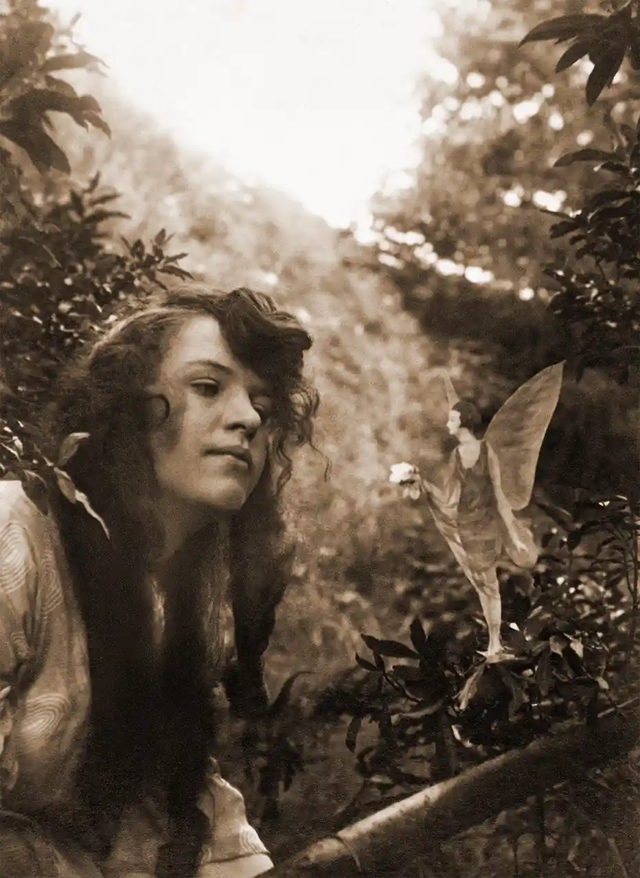 A sepia-toned vintage photograph shows a young woman with long, wavy hair looking pensively to the side, while a small fairy…