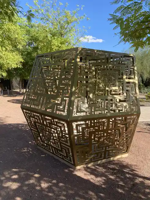 A large, cube-shaped outdoor sculpture with intricate maze patterns cut into its sides stands on a gravel path under a blue…