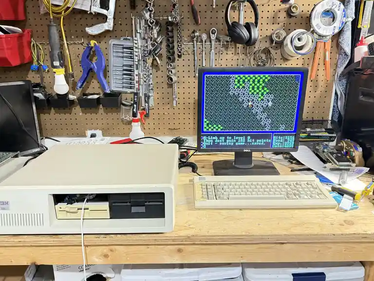 A beige IBM PC from the 1980s sits on a cluttered wooden workbench, connected to a modern Dell monitor displaying a…