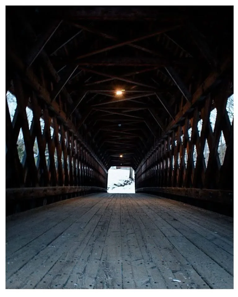 A low-angle shot looking down a long, dark wooden covered bridge with exposed beams and lattice sides, leading to a bright…
