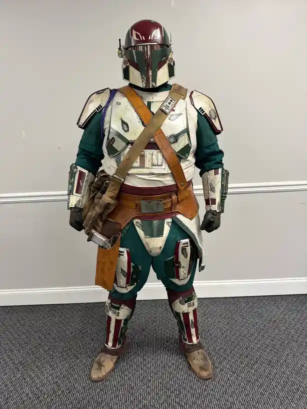 A person in a full-body, highly detailed Boba Fett cosplay stands facing forward against a plain wall. The costume features…