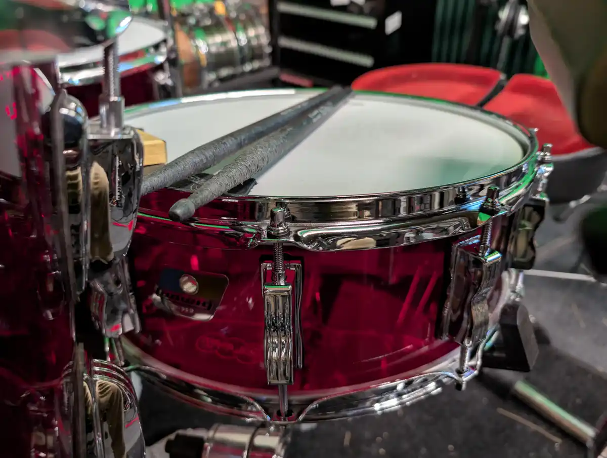 A close-up photo of a shiny red snare drum with chrome hardware, two drumsticks resting across the drumhead, and a small…