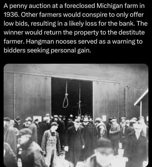 A black-and-white historical photograph shows a crowd of men in 1930s attire gathered outside a wooden barn. Two hangman's…