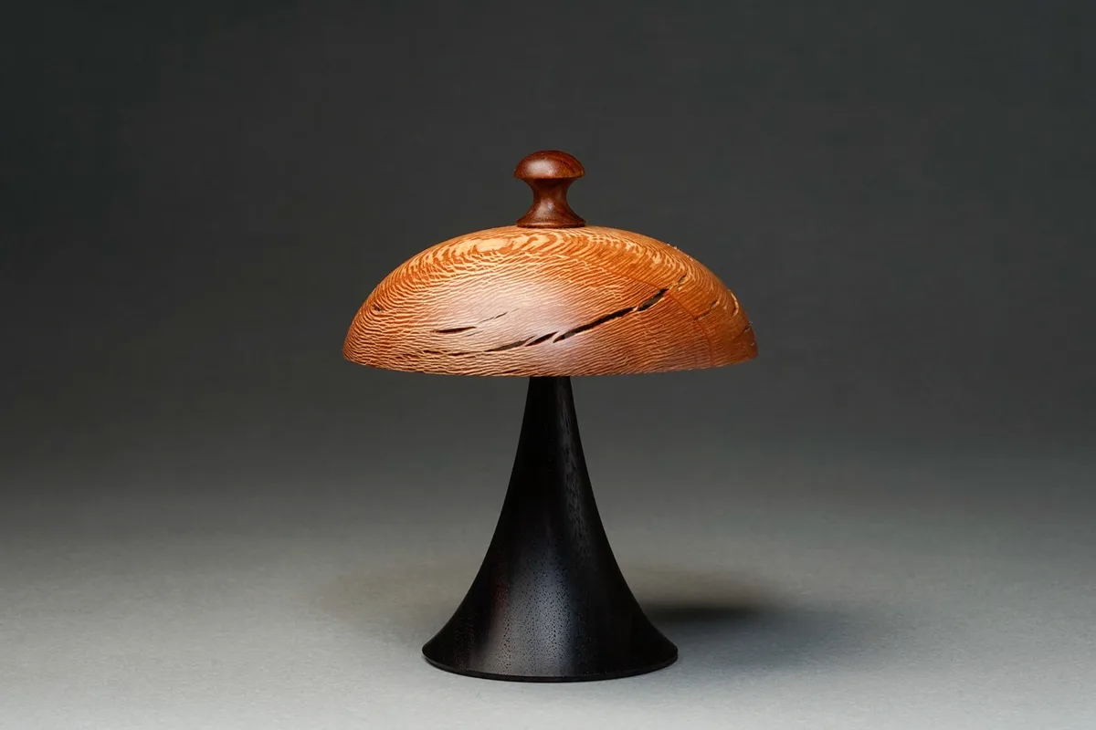 A wooden mushroom-shaped sculpture with a domed cap featuring carved lines and a small knob on top, mounted on a black…