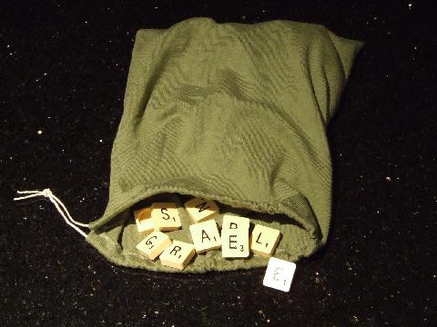 A green drawstring bag is open, spilling out Scrabble letter tiles onto a dark, speckled surface. Visible tiles spell 'S'…