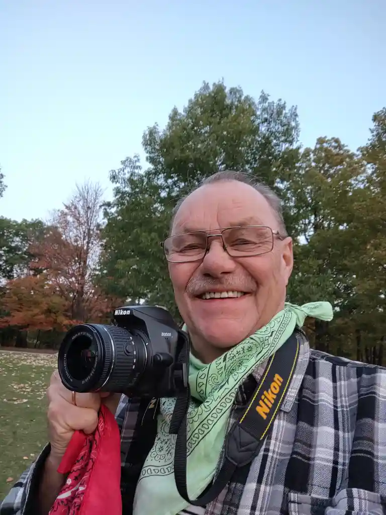 A cheerful older man with glasses and a green bandana smiles while holding a Nikon D3500 DSLR camera. He wears a plaid shirt…