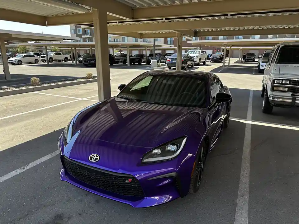 A shiny purple Toyota GR Supra sports car is parked in a covered parking lot, with other vehicles visible in the background…
