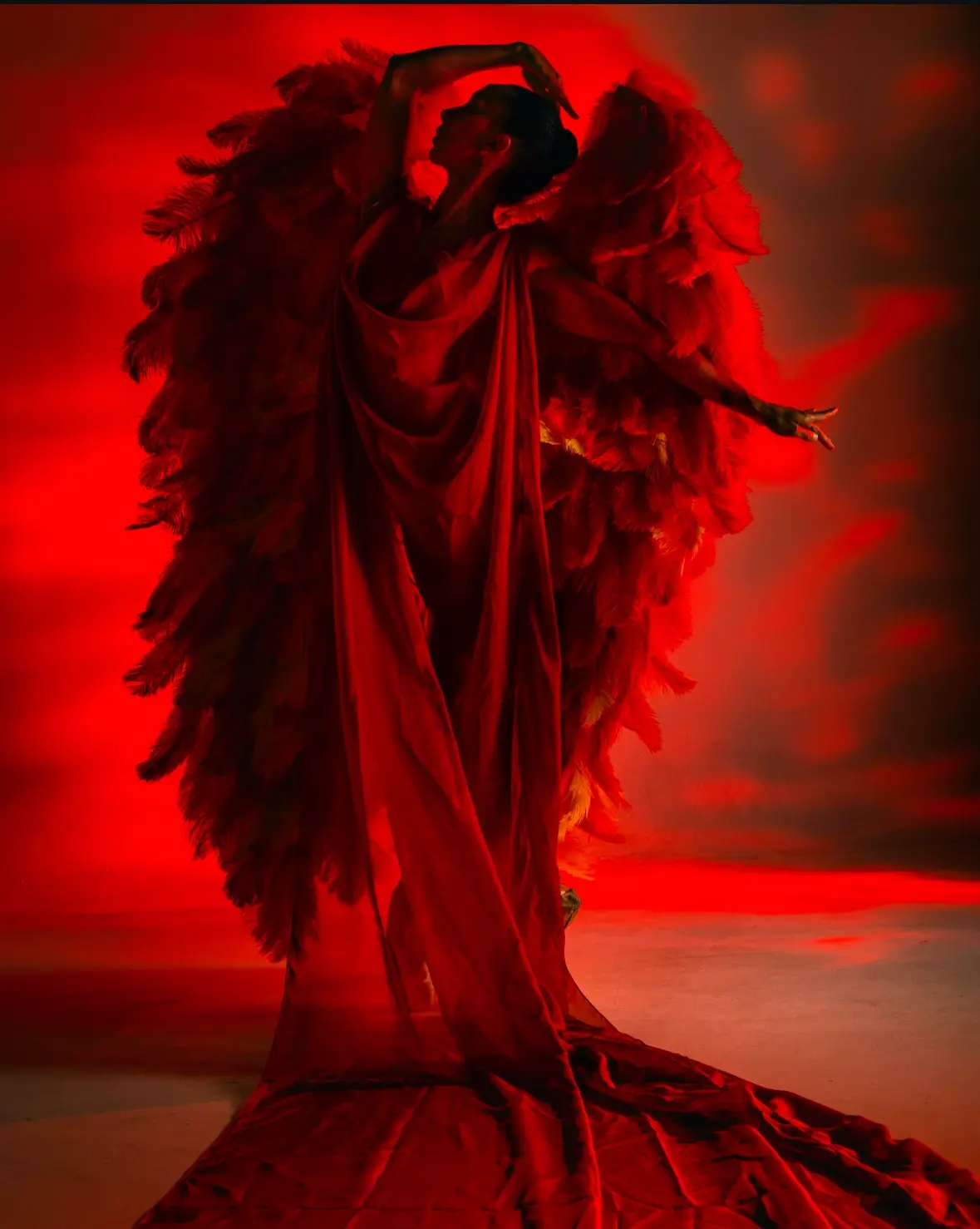 A person in a dramatic red costume with large feathered wings poses against a red-lit background, looking upward with one…