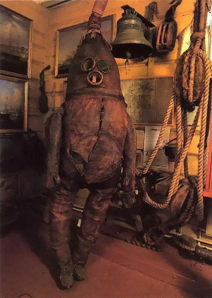 A vintage, heavily worn, full-body diving suit with a large helmet and glass eye pieces stands upright in a dimly lit room…