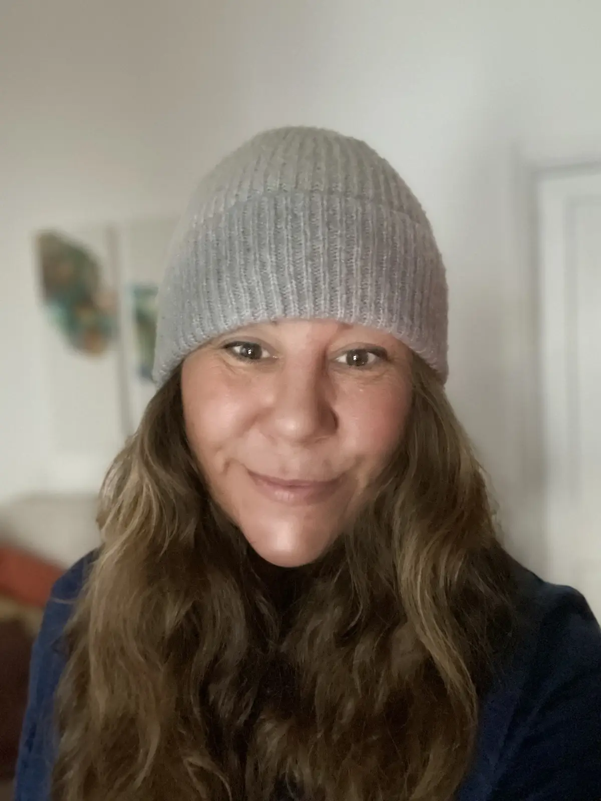 A woman with long brown hair and a light gray knit beanie smiles at the camera while wearing a dark blue top. The background…
