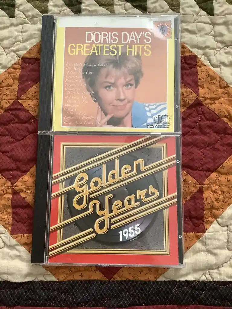 Two CD cases are stacked vertically on a quilted fabric background. The top case is for 'Doris Day's Greatest Hits'…
