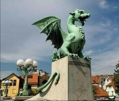 A large, green, weathered bronze dragon statue with wings spread, perched atop a stone pedestal in an urban setting. The…