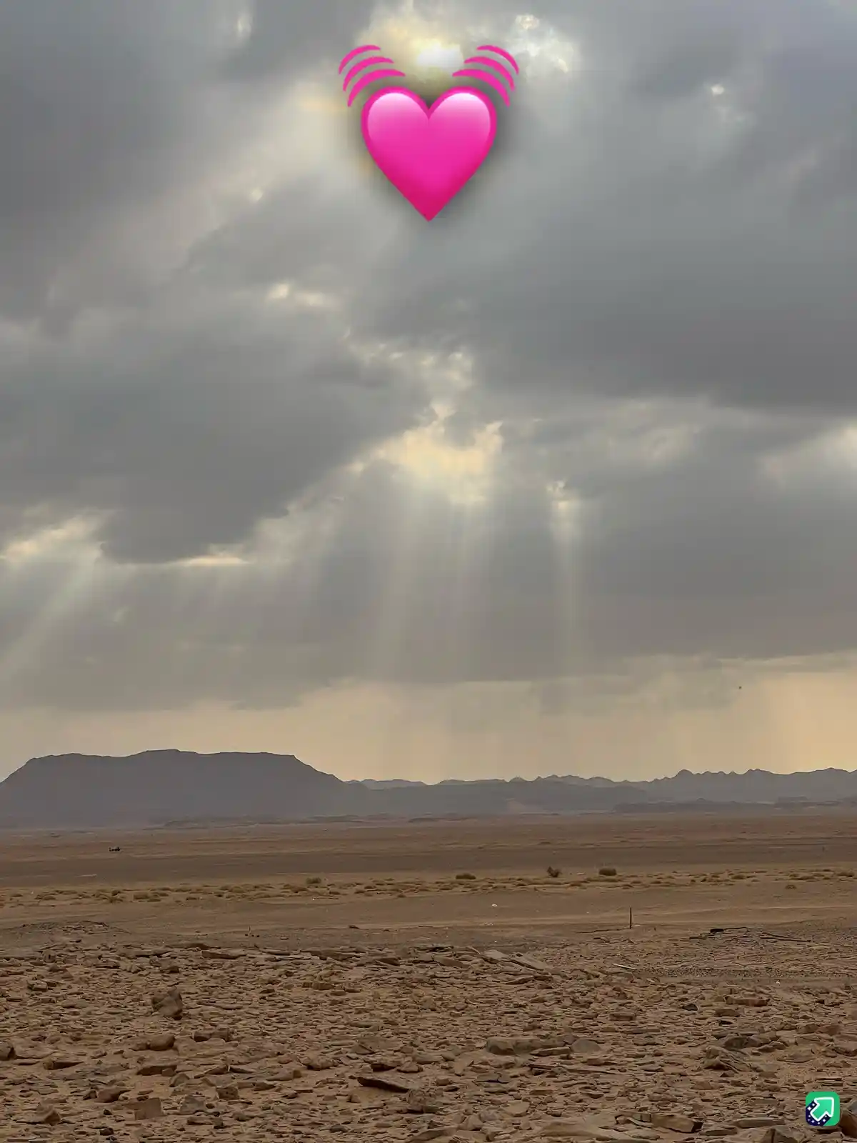 A wide shot of a barren desert landscape under a cloudy sky with dramatic sunbeams breaking through. A bright pink heart…