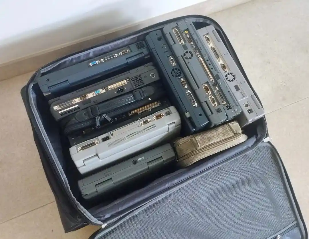 An open black suitcase filled with multiple old, bulky laptops and portable devices, all stacked with their ports and vents…