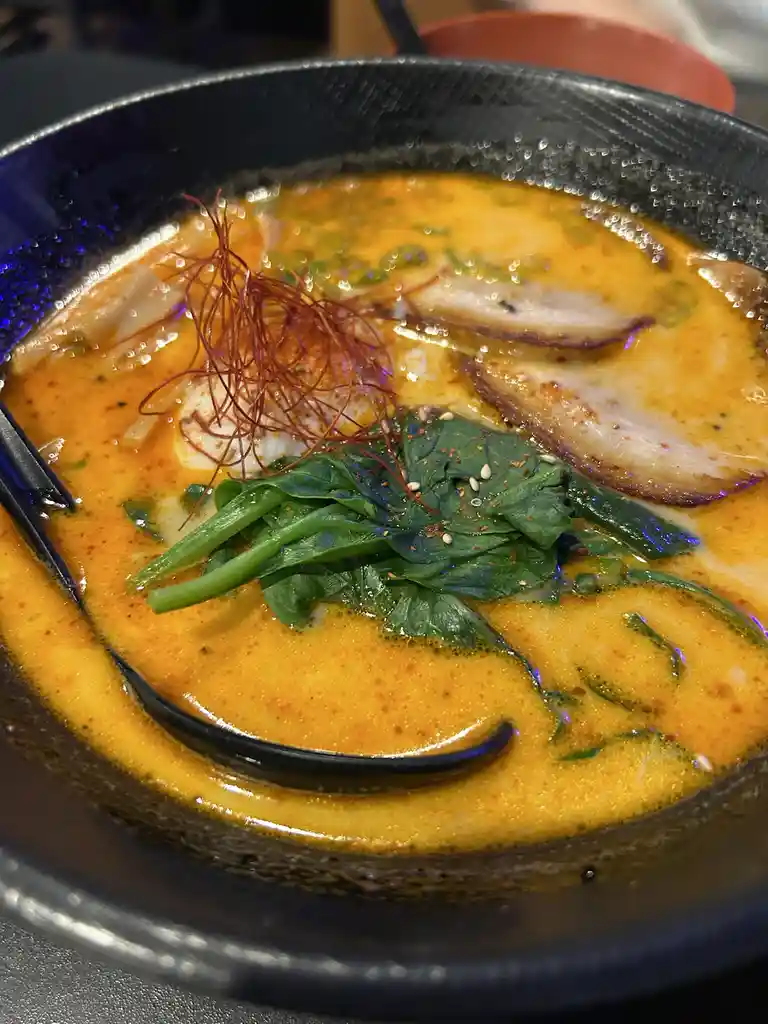 a close-up photo of a bowl of ramen with a creamy orange broth, topped with spinach, sesame seeds, crispy fried noodles, and…