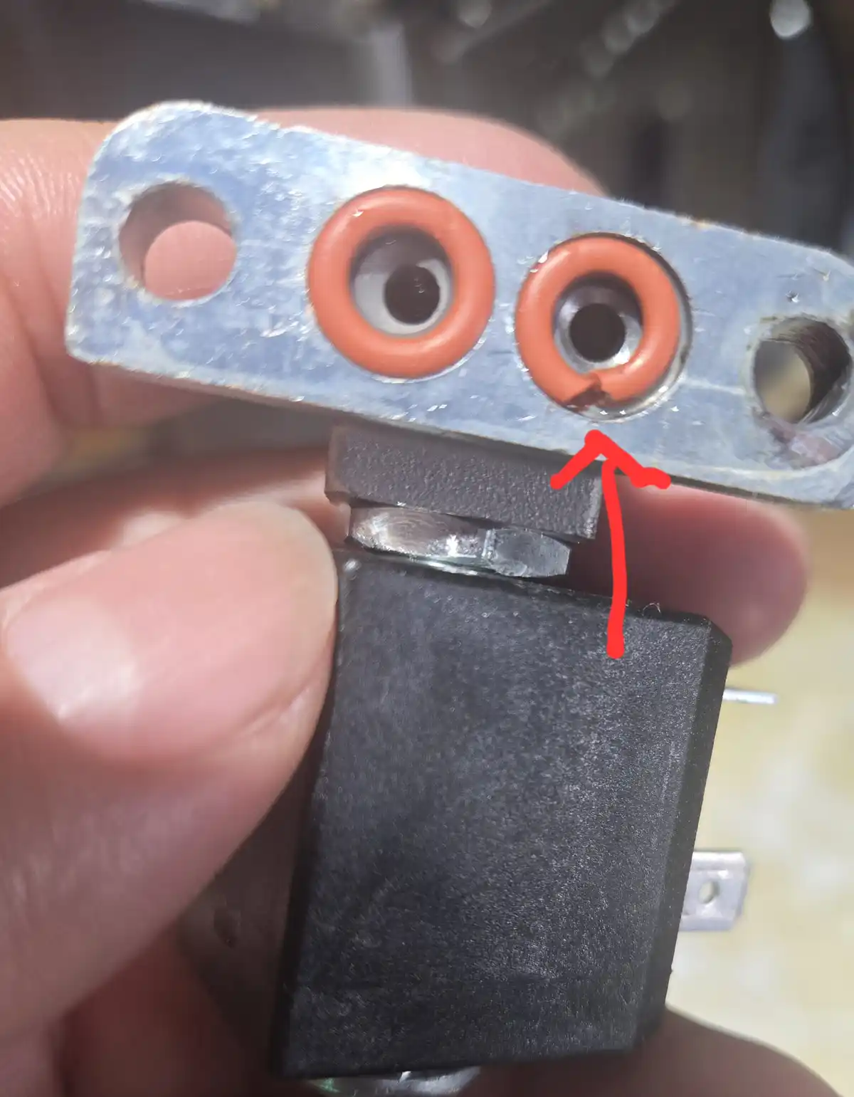 A close-up photo shows a hand holding a metal bracket with two orange rubber O-rings that resemble eyes, and a black…