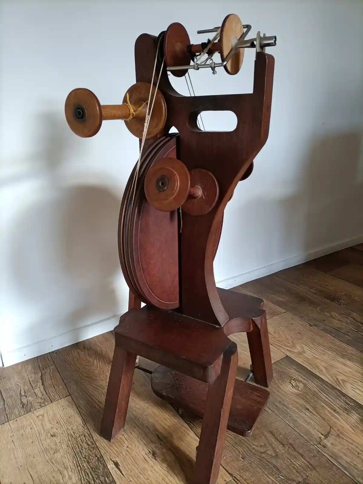 A wooden spinning wheel with multiple spindles and a seat, standing on a wooden floor against a white wall, overlaid with…