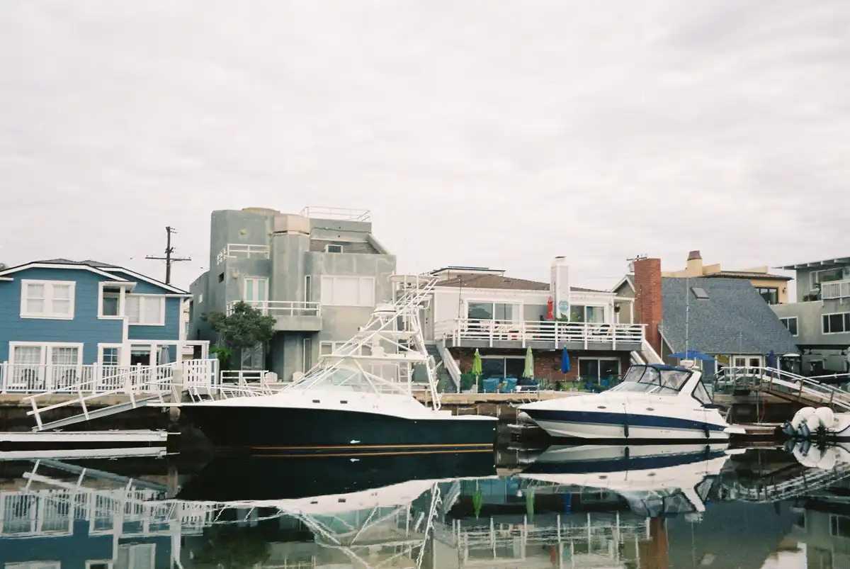 A calm harbor scene with several boats docked in front of colorful waterfront houses under an overcast sky. The water…