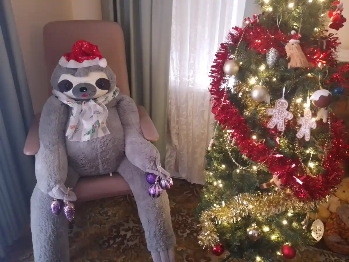 A large grey stuffed sloth plushie wearing a red and white Santa hat and a patterned scarf sits in a pink armchair next to a…