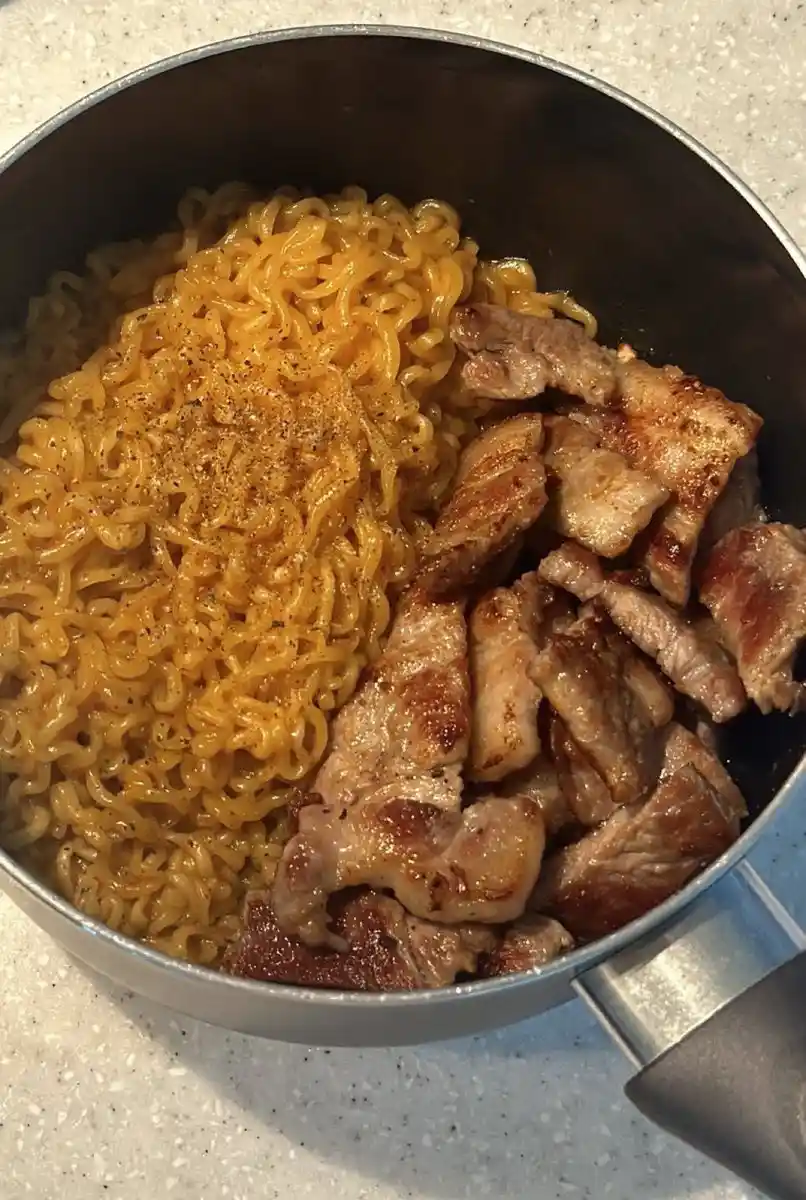 a close-up photo of a metal pot containing cooked ramen noodles seasoned with black pepper, alongside a pile of seared…