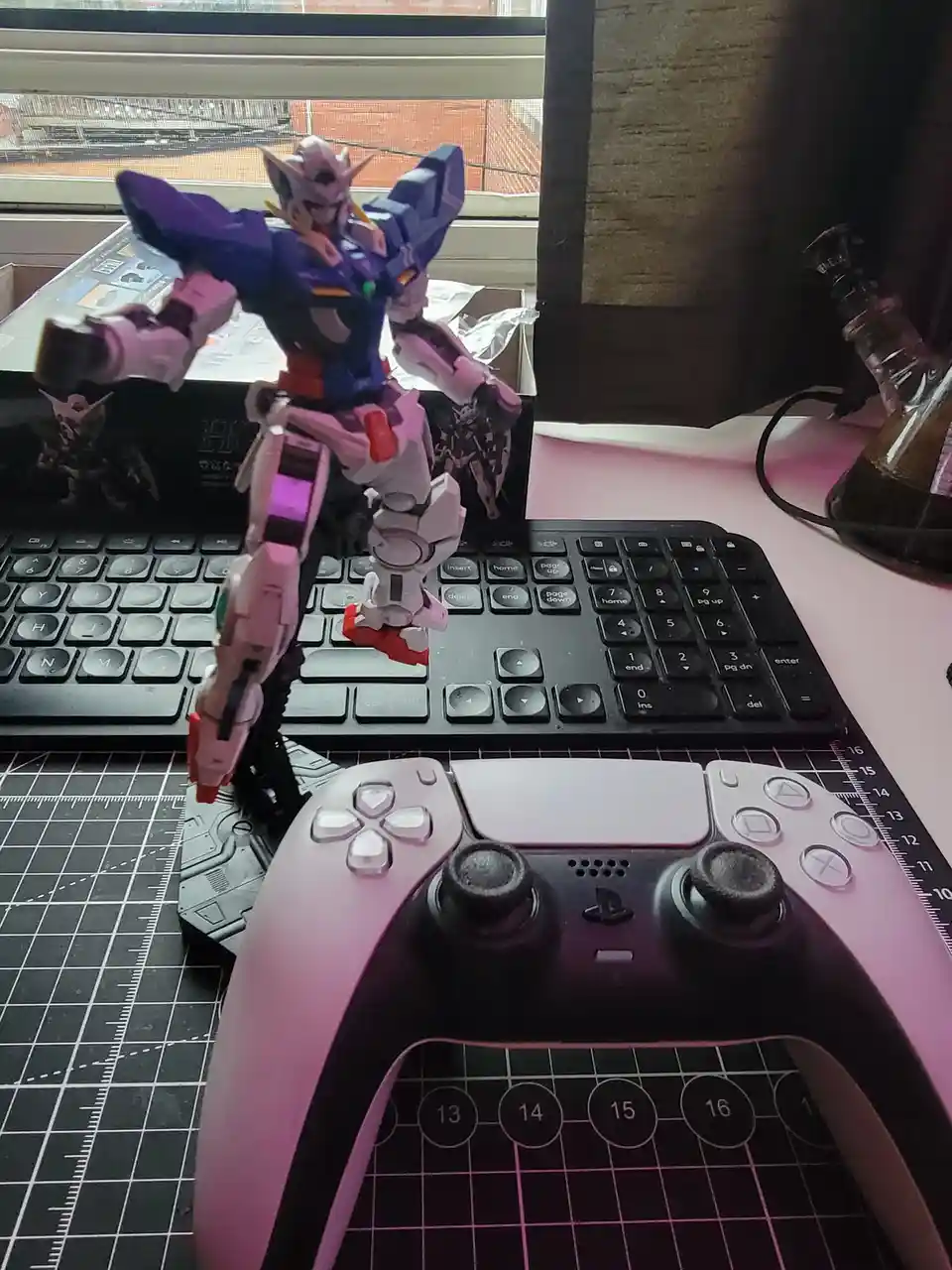 A purple and white Gundam model stands on a black keyboard, with a pink and black PlayStation 5 controller in the…