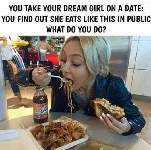 A woman with blonde hair is seated at a restaurant table, enthusiastically eating noodles from a takeout container with a…
