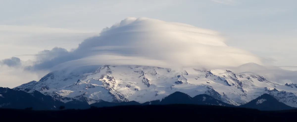 A snow-capped mountain is topped by a large, smooth, elongated cloud that visually resembles the roof of a car, creating a…