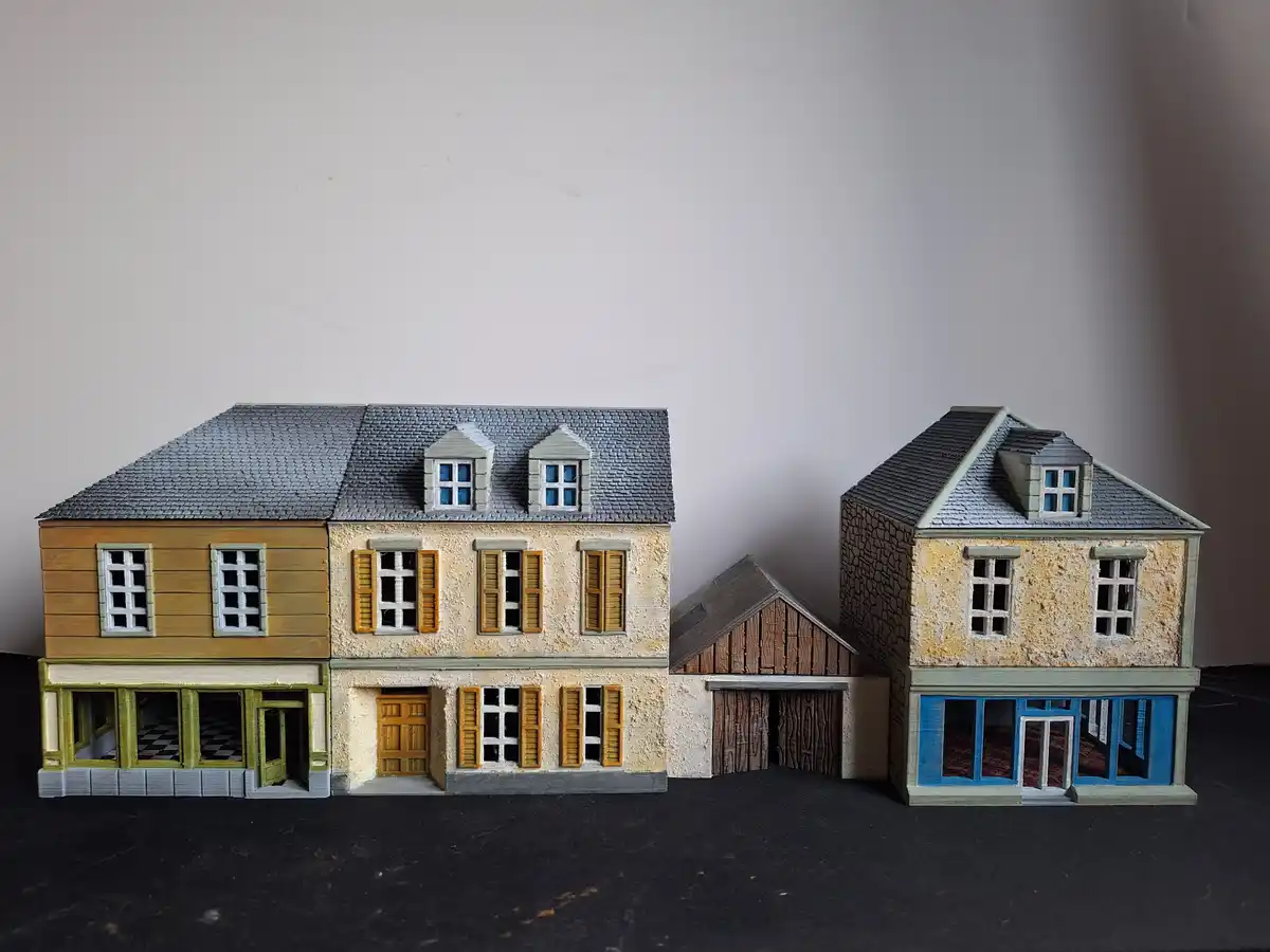 The image displays two 3d-printed miniature European-style houses with gray roofs, shuttered windows, and detailed facades…
