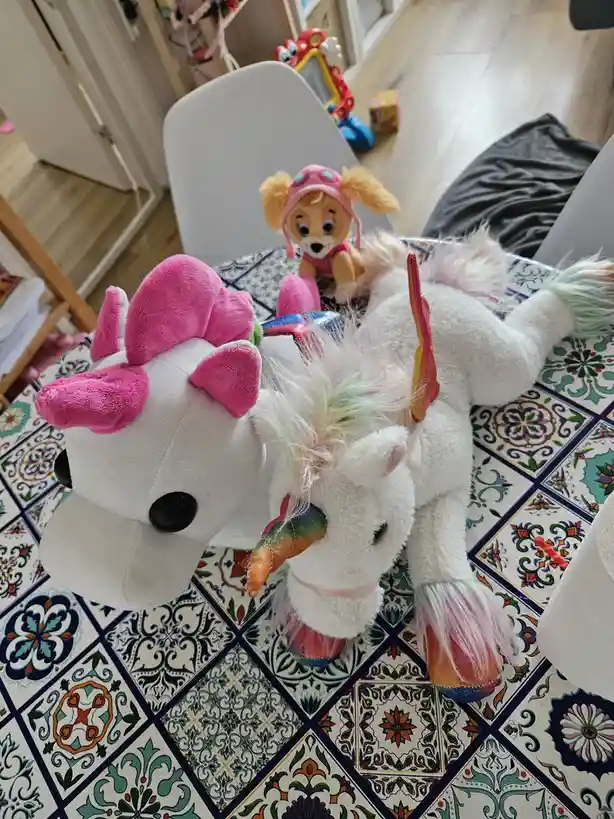 A plush white unicorn toy with a pink hat and rainbow horn lies on a colorful tiled table, with a small stuffed dog wearing…