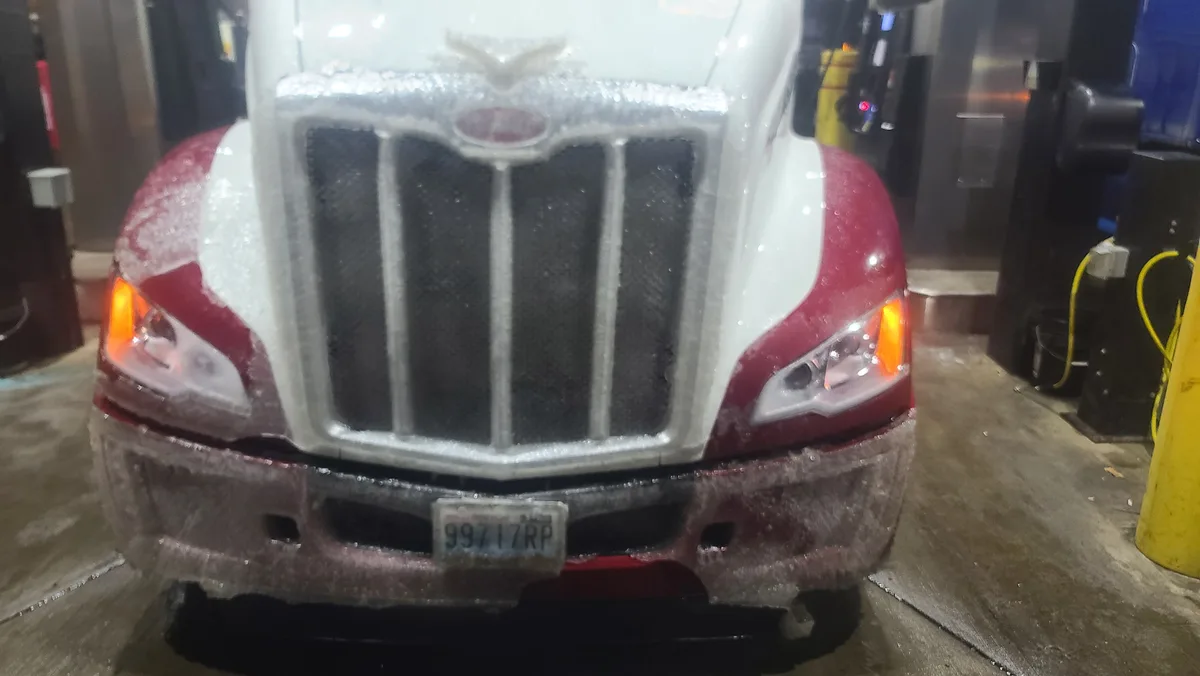 A red and white semi-truck is covered in ice and snow, particularly on its front grille, hood, and bumper, while its…