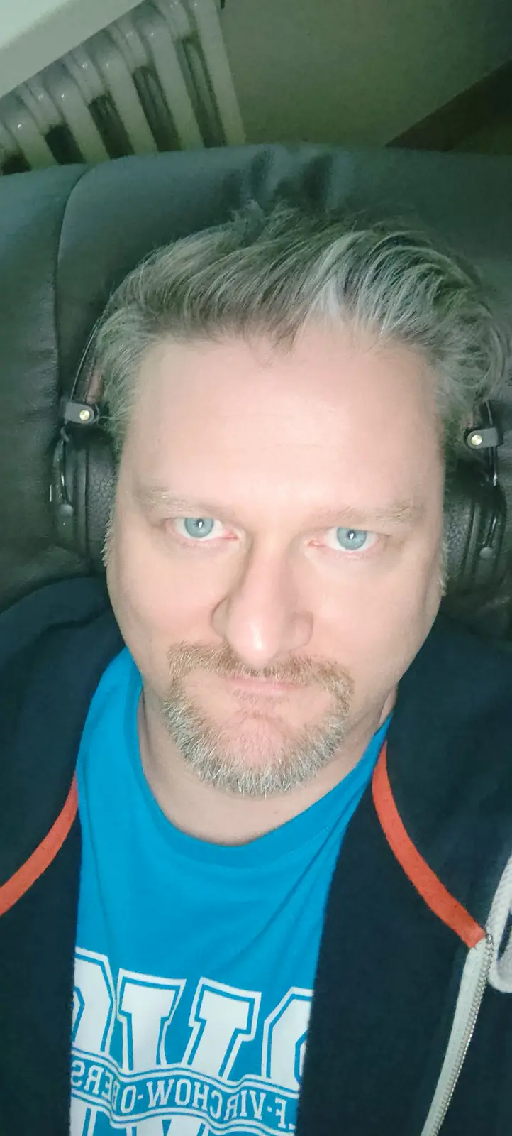 A man with gray hair and blue eyes, wearing headphones, a blue t-shirt, and a dark hoodie, stares intensely at the camera…