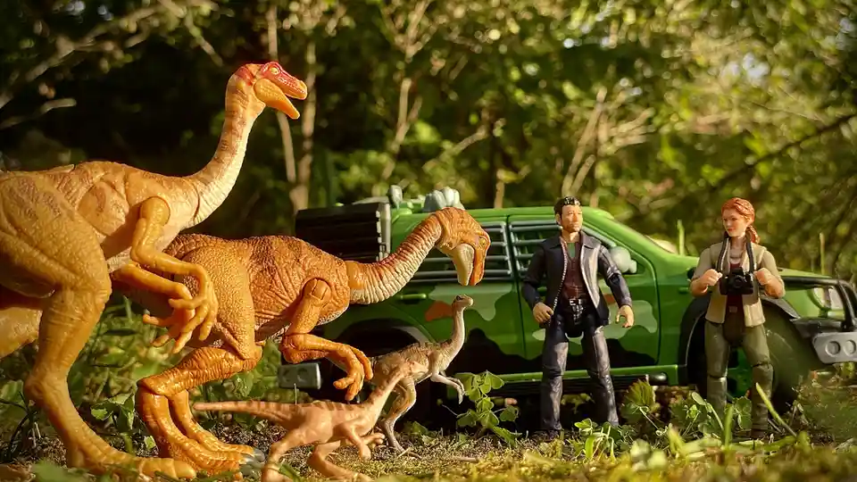 A staged photo of toy dinosaurs and human figures in a forest setting, with a green toy truck in the background. Two large…