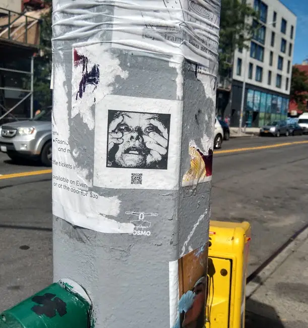 A weathered urban pole covered in peeling flyers and stickers, one of which features a black-and-white distressed portrait…