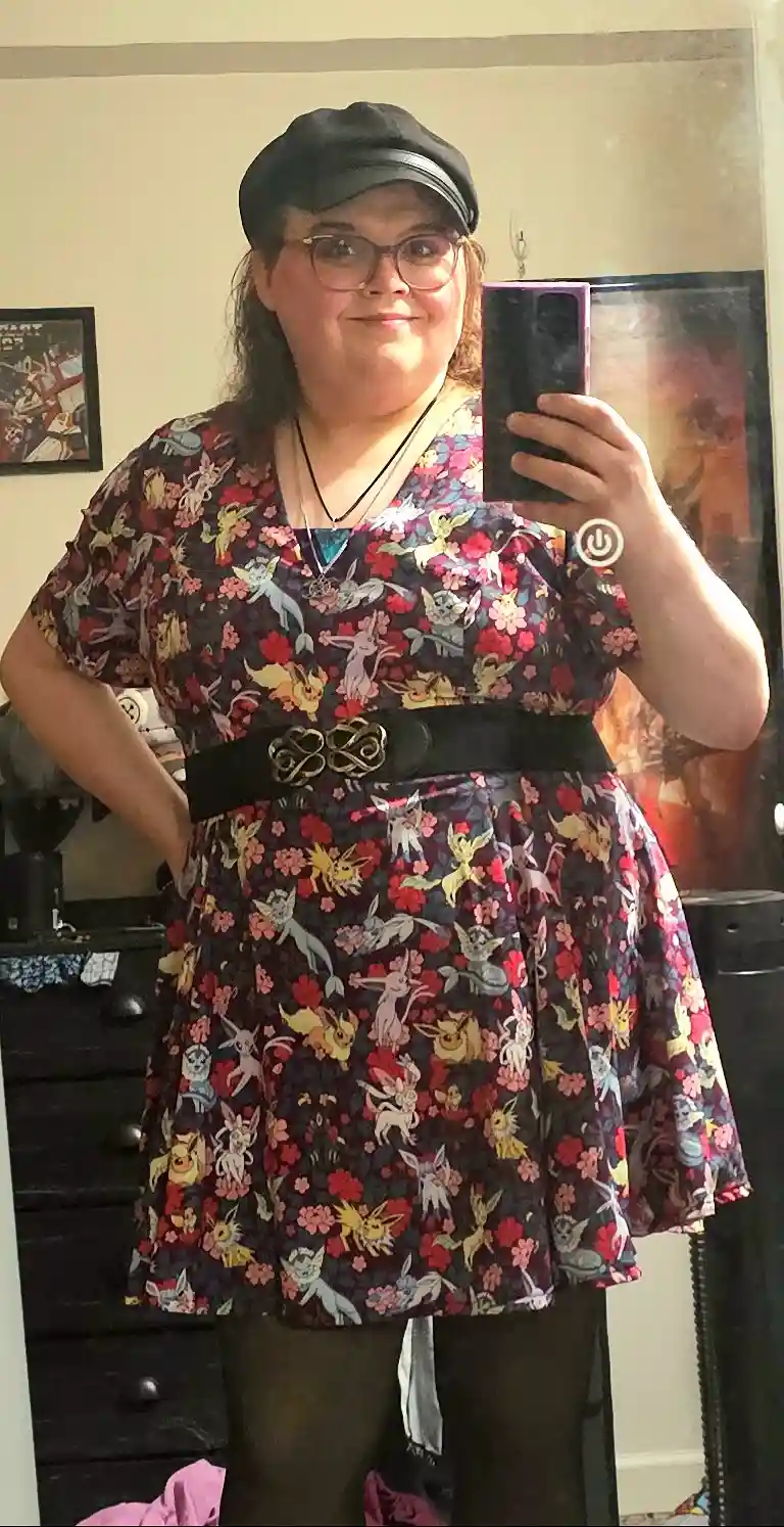 A person wearing a black cap and glasses takes a mirror selfie while holding a phone. They are wearing a dark floral dress…