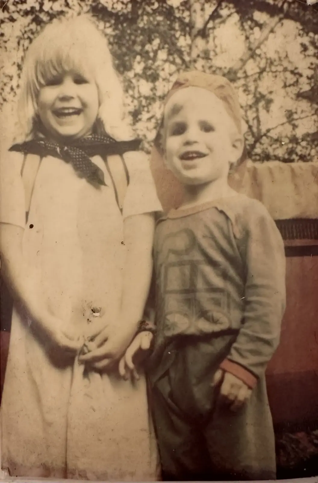 A sepia-toned, vintage photograph shows two young children standing side by side outdoors, smiling broadly. The child on the…