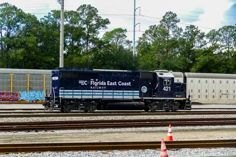 A Florida East Coast Railway locomotive numbered 421 is shown on train tracks with trees and overhead power lines in the…