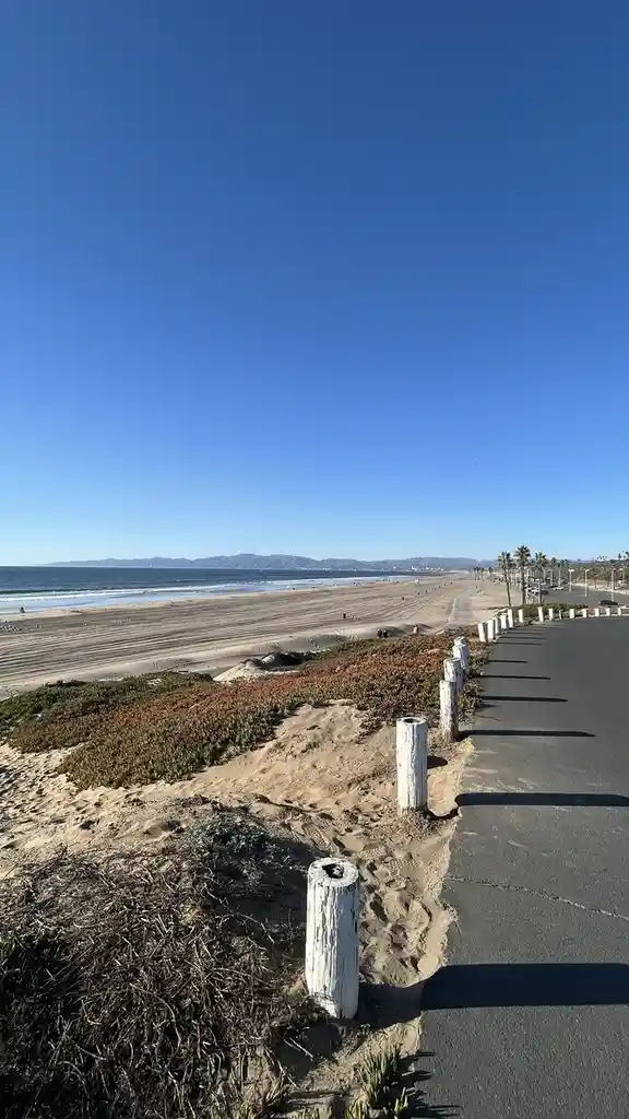 A scenic beach view with a clear blue sky, ocean waves, and a paved path lined with weathered white posts. The overlaid text…
