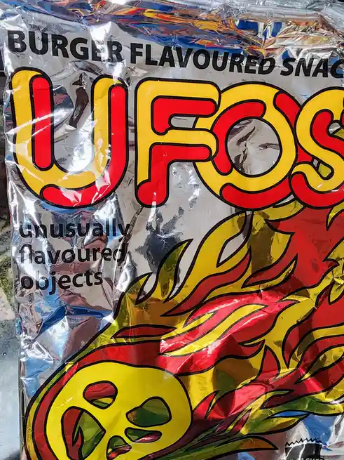 A close-up of a crinkled, shiny silver snack bag with bold red and yellow text reading 'UFOs' and 'BURGER FLAVOURED SNACK'…