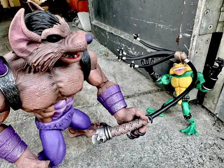 Two action figures are staged in a gritty outdoor setting: a large, muscular, pink-skinned, horned humanoid with purple…
