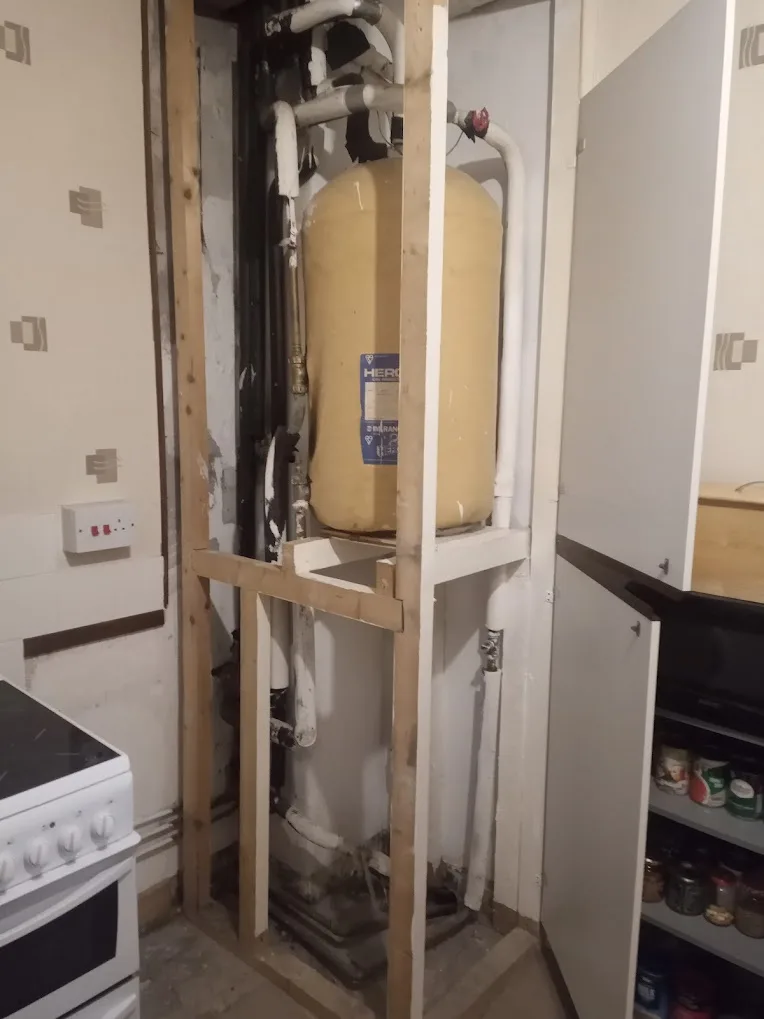 A kitchen corner showing exposed wooden framing around a large yellow water heater tank with pipes, next to a white stove…