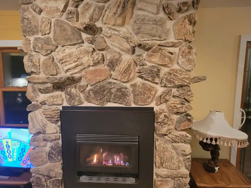 A cozy room features a stone fireplace with a lit gas fire, next to a lamp and a TV showing a slot machine game. The…