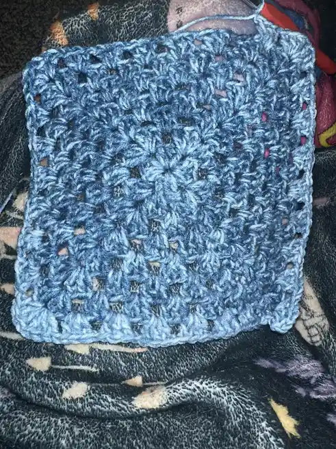 A close-up photo shows a handmade, light blue crocheted square with a textured, floral-like pattern, resting on a dark…