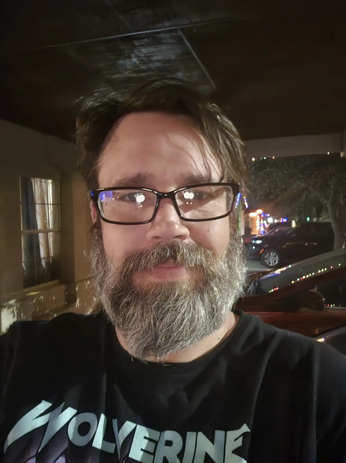 A man with a graying beard and glasses takes a selfie at night, wearing a black t-shirt with the word 'WOLVERINE' in large…