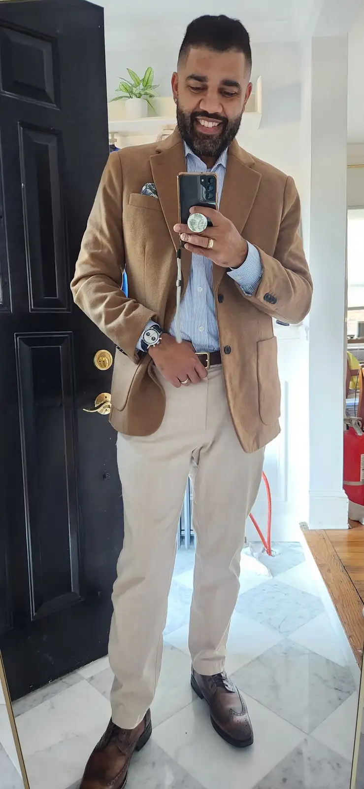 A man with a beard and short hair takes a mirror selfie in a stylish camel blazer, light-colored pants, and brown brogues…