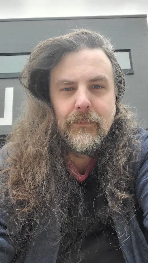 A man with long, wavy brown and gray hair and a full beard stares directly at the camera with a neutral expression. He wears…