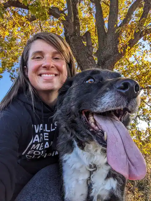 A woman with freckles and a black hoodie smiles while holding a large black and white dog with its tongue out, set against a…
