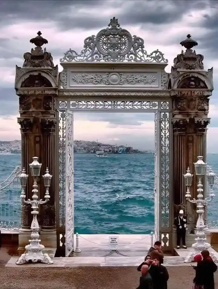 An ornate, white, baroque-style metal archway frames a view of the sea and distant cityscape under a cloudy sky. People are…